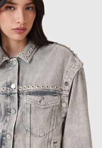 Giacca in denim grigio sbiadito con bottoni a pressione in metallo e dettagli con occhielli sulle spalle, dotata di una tasca sul petto e una texture consumata.