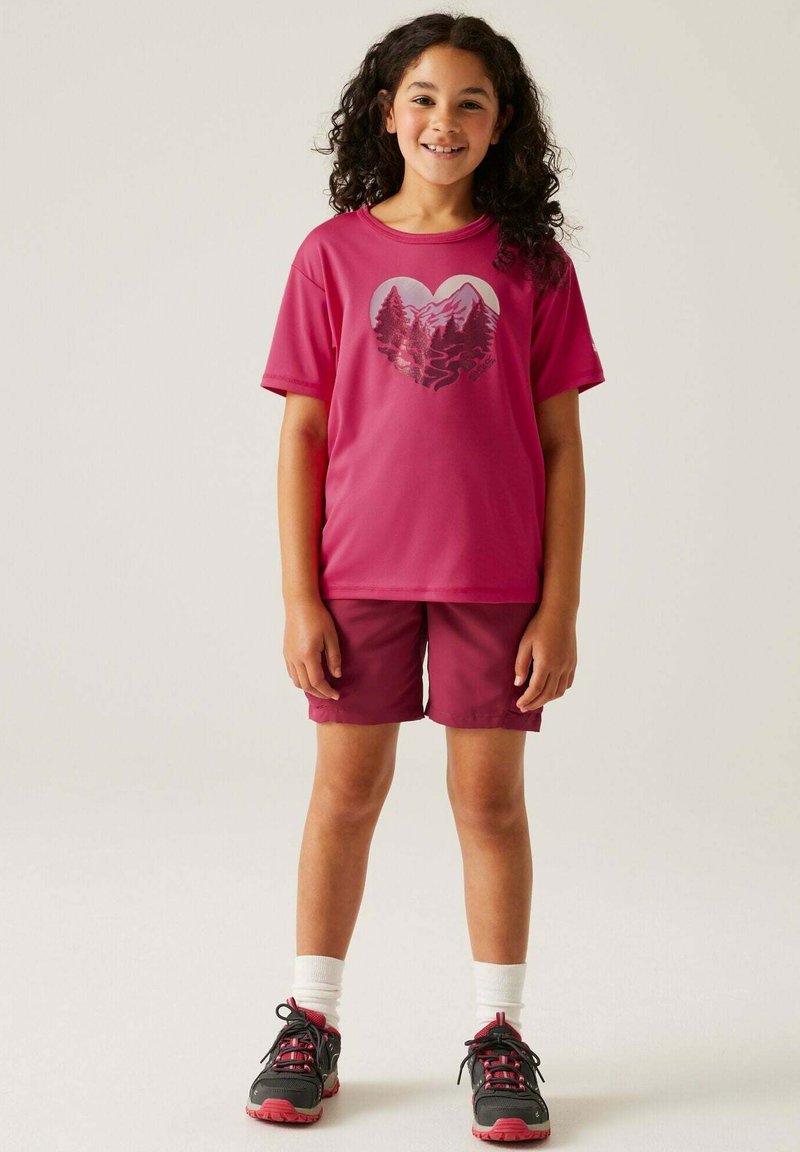 Camisa deportiva rosa con un gráfico de montaña, combinada con pantalones cortos burdeos. Lleva zapatillas negras y rosas con calcetines blancos. Textura de tela suave.