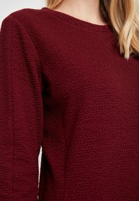 Burgundský texturovaný top s kulatým výstřihem a dlouhými rukávy, se zvrásněným vzorem látky.