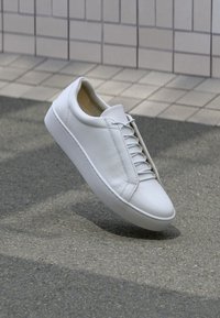 Zapatilla blanca de piel con puntera redonda, suela plana y seis pares de ojetes. Textura suave y diseño minimalista con detalles de costura.