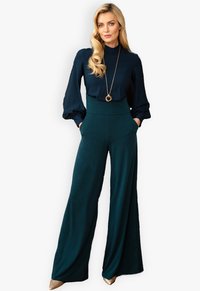Navybluse mit Puffärmeln, hochgeschnittene grüne weit geschnittene Hose mit zwei Seitentaschen, akzentuiert durch eine lange goldene Halskette und glitzernde Schuhe.