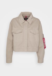 Beige cropped Jeansjacke mit langen Ärmeln, vorderer Knopfverschluss, zwei Brusttaschen und einem roten Etikett-Akzent am Ärmel.