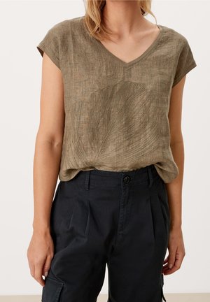 Vrouw draagt een olijfgroen kort mouw V-hals blouse met bladborduursel, ingestopt in een zwarte, hooggetailleerde cargobroek.