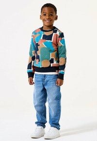 Un ragazzo giovane sorridente in piedi, che indossa un maglione a motivo geometrico multicolore, jeans azzurri chiari e sneakers bianche su uno sfondo bianco.