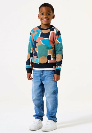 Un ragazzo giovane sorridente in piedi, che indossa un maglione a motivo geometrico multicolore, jeans azzurri chiari e sneakers bianche su uno sfondo bianco.