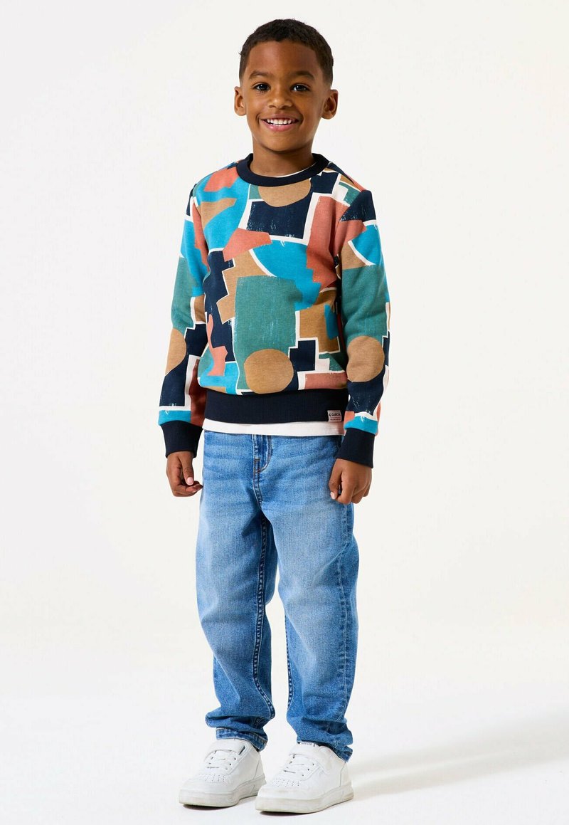 Un ragazzo giovane sorridente in piedi, che indossa un maglione a motivo geometrico multicolore, jeans azzurri chiari e sneakers bianche su uno sfondo bianco.