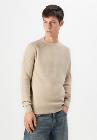 Giovane uomo con capelli corti e scuri, che indossa un maglione beige a trama e jeans blu, mentre guarda di lato con una mano in tasca.