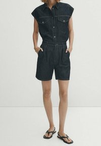 Chemise en denim foncé à manches courtes avec col pointu, poches poitrine doubles et short taille haute assorti. Portée avec des tongs noires.