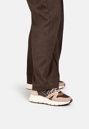 Person trägt braune Hose und beige, braune und schwarze Sneaker mit weißen Schnürsenkeln, steht auf einem hellgrauen Boden.