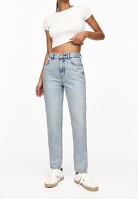 Kvinna i ljusblå högmidjade jeans, vit kortärmad topp och vita sneakers med beige sulor, står framför en vit bakgrund.