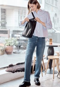Jeune femme en chemise rayée et jeans souriant en regardant à l'intérieur d'un sac à bandoulière noir dans un café moderne.
