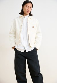 Light cream jacket with a zipper and front pocket, worn over a white and blue striped shirt, paired with black trousers. Simple design.