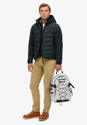 Hombre joven con chaqueta acolchada oscura, pantalones beige, zapatos marrones, sosteniendo una mochila blanca con detalles y logo en negro en la mano derecha.