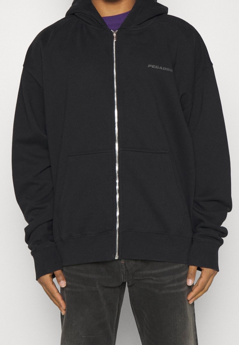 Person som bär en svart zip-up hoodie med framfickor och "PEGADOR"-logotyp på bröstet, ihop med mörkgrå byxor.