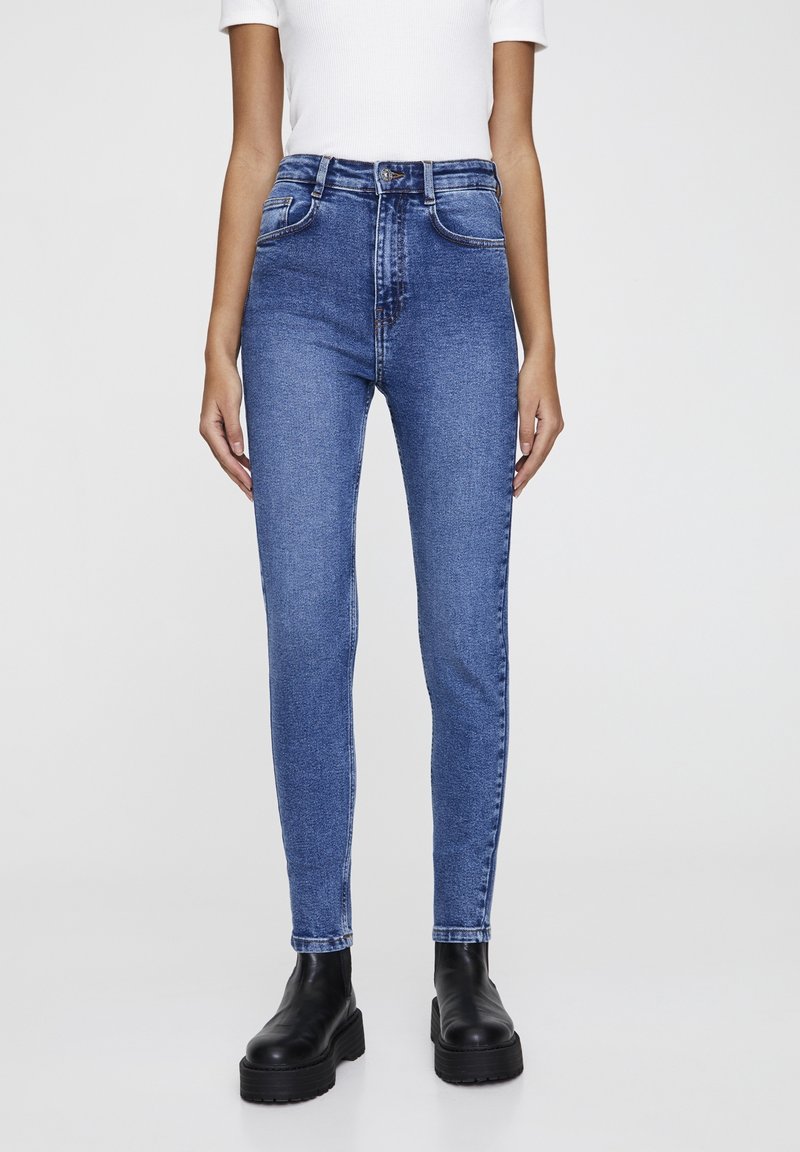 Personne portant un jean skinny bleu taille haute, un haut blanc à manches courtes et des bottines noires épaisses, debout devant un fond uni.