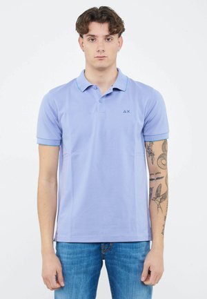 Jonge man met kort bruin haar, gekleed in een lichtpaarse polo en blauwe spijkerbroek, met tatoeages zichtbaar op zijn rechteronderarm.