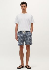 Tenue décontractée pour homme composée d'un t-shirt blanc uni et d'un short de bain bleu marine à motifs avec un cordon de serrage, complétée par des tongs noires.