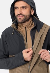 Kurtka typu hoodie z czarną górną częścią i brązową dolną częścią, wykonana z teksturowanej tkaniny, z zamkami błyskawicznymi i regulowanymi sznureczkami.
