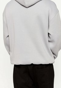 Šedá mikina s kapucí a zadním švem, která má volný střih a texturovanou látku. V kombinaci s černými kalhotami s dvěma zadními kapsami.