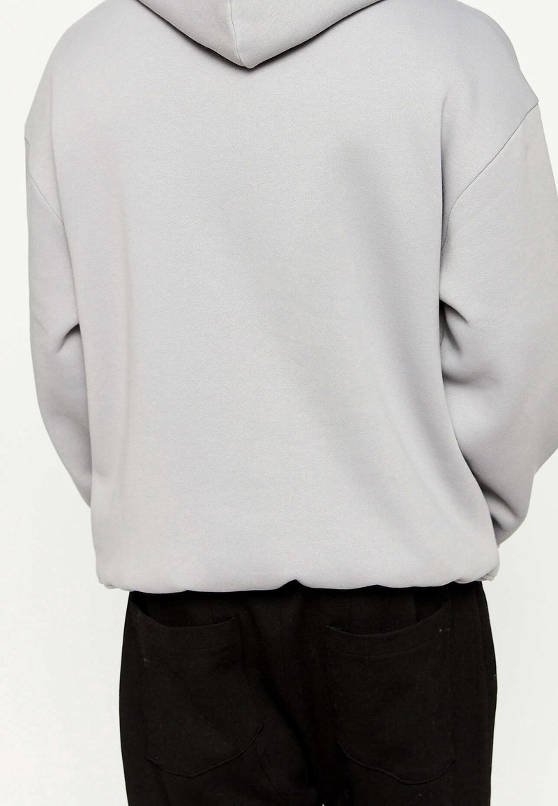 Šedá mikina s kapucí a zadním švem, která má volný střih a texturovanou látku. V kombinaci s černými kalhotami s dvěma zadními kapsami.