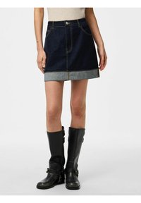 Gonna di jeans blu scuro con orlo blu chiaro. Presenta tasche frontali e chiusura con bottone. Indossata con stivali neri fino al ginocchio.