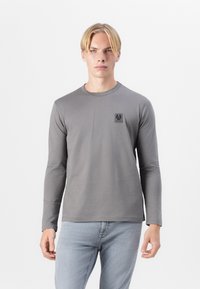 Tricou gri cu mâneci lungi din material moale, cu un mic patch cu logo negru pe piept, guler rotund și design ajustat.