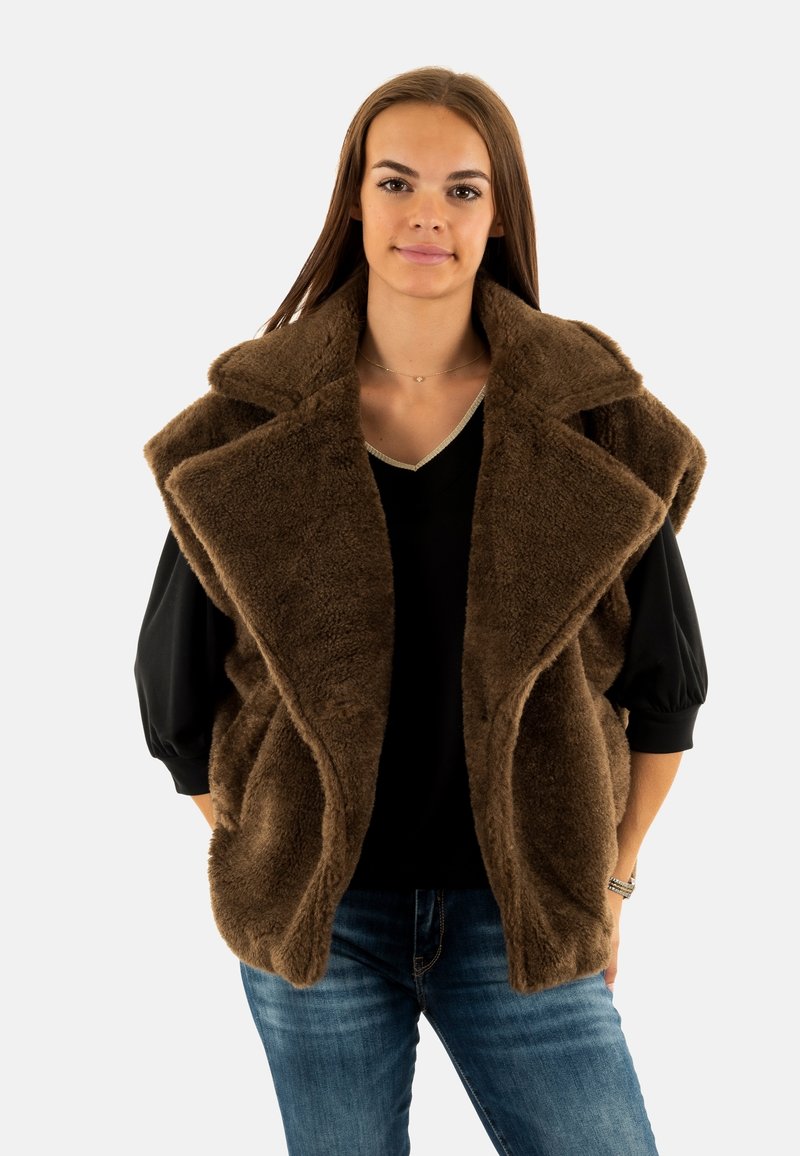 Bruine nepbont gilet met een brede kraag, een open voorkant en twee zijzakken. Draag het over een zwarte top, gecombineerd met blauwe spijkerbroeken.