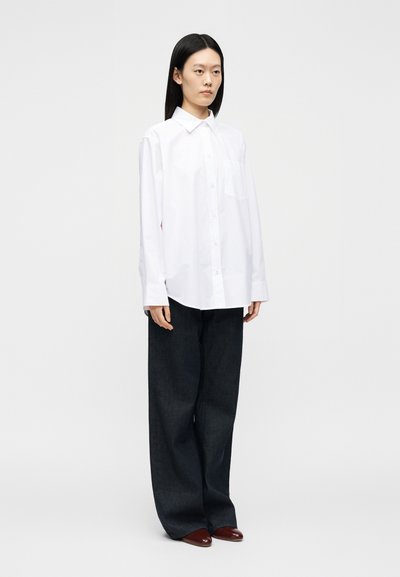 Femme aux cheveux noirs raides portant une chemise blanche ample à boutons, un pantalon noir large et des chaussures marron pointues, debout devant un fond uni.