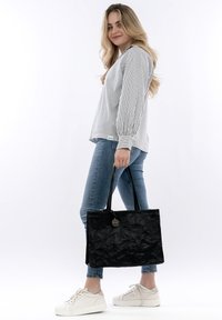 Borsa a tote nera con texture, dotata di due manici lunghi e un'etichetta circolare. Il modello indossa una maglietta a righe e jeans blu, con sneakers bianche.