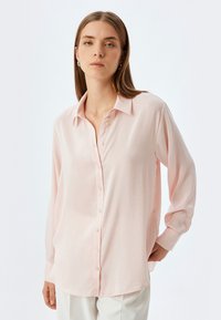 Camisa de manga larga de color rosa claro con cierre de botones en la parte delantera, cuello clásico y un ajuste ligeramente holgado. Textura suave y un ligero brillo.