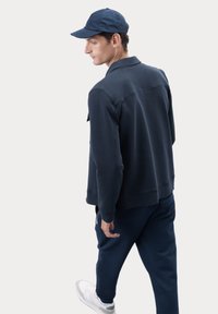 Felpa navale con design a colletto, tessuto testurizzato e polsini a coste, abbinata a pantaloni coordinati e scarpe da ginnastica bianche.