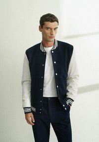 Giacca varsity blu navy e crema con polsini a righe, bottoni a pressione e una maglietta bianca a girocollo sotto, abbinata a pantaloni scuri.