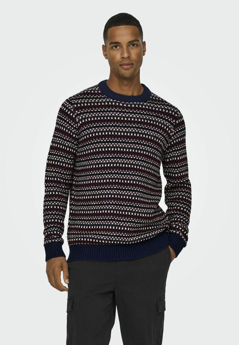 Pull en maille navy avec un motif blanc et bordeaux, manches et col côtelés. Le tissu a une apparence texturée et confortable.