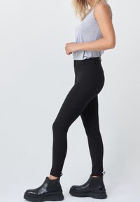 Černé legíny s jemnou texturou, vysokým pasem a postranními kapsami. V kombinaci s černými kotníkovými botami a šedým topem bez rukávů.
