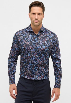 Mann trägt ein enges, langärmliges Hemd mit einem dunklen Paisley- und Blumenmuster in Blau- und Rottönen, kombiniert mit dunklen Hosen.