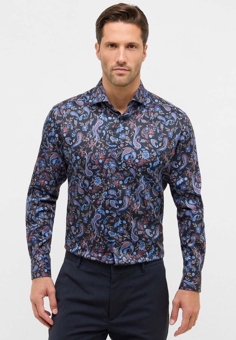 Man in een fitted, langemouwen shirt met een donkere paisley- en bloemprint in blauw en rode tinten, gecombineerd met donkere broek.