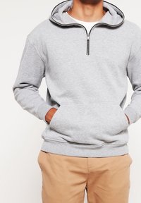 Uomo che indossa una felpa grigia chiara con mezzo zip e tasca frontale, pantaloni beige, mani in tasca, in piedi contro uno sfondo neutro.