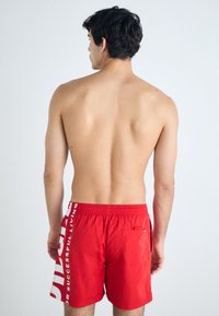 Short de bain rouge en matériau léger, doté d'une taille élastique et d'une poche latérale avec un texte graphique blanc sur le côté gauche.