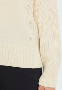 Strikkegenser i kremfarge, med ribbestrikket kant og mansjetter, samt et teksturert stikkmønster på kroppen. Sidestykke detaljer gir dimensjon.