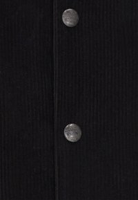 Sort corduroy stof med lodrette ribber, prydet med en sølvknap mærket "Dreimaster." Teksturen ser blød og tæt ud.