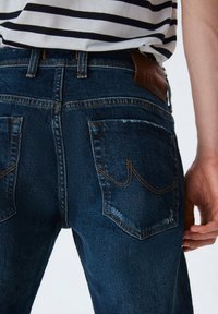 Los jeans de mezclilla azul oscuro tienen una silueta ajustada, un sutil desgastado y un parche de cuero marrón en contraste. Las costuras delinean los bolsillos traseros.