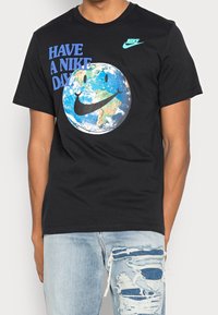 Svart bomullströja med en grafik av jorden med ett leende ansikte och Nike-swoosh, samt texten "HA EN NIKE-DAG" i blått.
