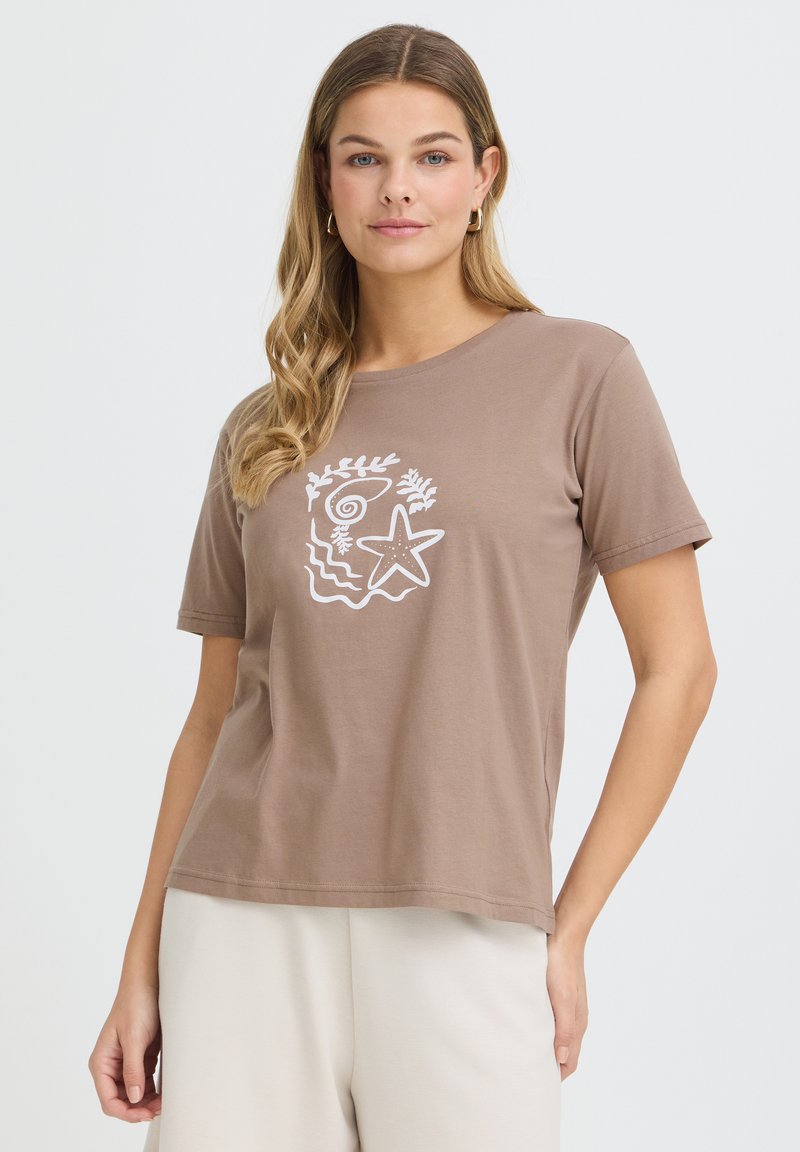 Femme aux longs cheveux ondulés portant des boucles d'oreilles créoles dorées, un t-shirt marron avec des motifs marins blancs, et un pantalon blanc sur un fond uni.