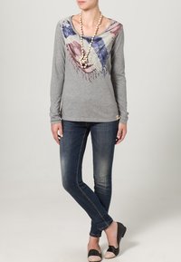 Graues Langarmoberteil mit buntem abstraktem Druck am Ausschnitt, kombiniert mit dunklen Skinny-Jeans und schwarzen sowie beigen Ballerinas.