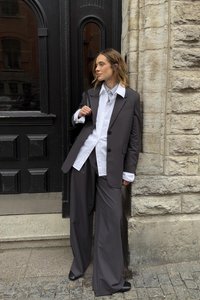 Pelēks pārmērīgi liels žakets un saskaņotas platleņķa bikses virs baltas krekli ar svītrotu apkakli. Gluda auduma, minimālisma dizains un smalkas detaļas.