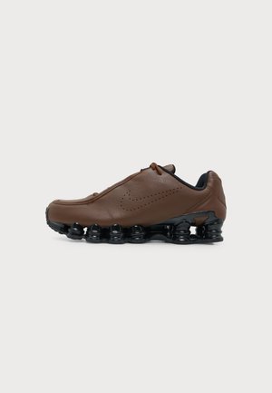 Chaussure de sport en cuir marron avec une semelle noire en forme de ressort et un swoosh Nike perforé sur le côté, montrée de profil sur fond blanc.