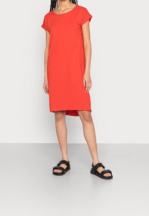 Femme portant une robe rouge ample jusqu'aux genoux avec des manches courtes retroussées et des sandales noires à bouts ouverts sur un fond uni.