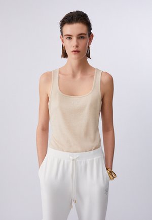 Donna che indossa un top lavorato a maglia beige senza maniche e pantaloni bianchi con coulisse, con braccialetti d'oro al polso, davanti a uno sfondo chiaro e semplice.
