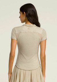Camicia attillata beige con scollatura rotonda, maniche corte e texture morbida. Presenta un logo sottile sulla parte posteriore e un orlo curvo.