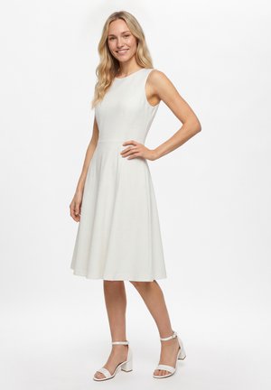 Glimlachende vrouw met lang blond haar draagt een mouwloze witte jurk tot net boven de knie en witte sandalen met blokhak, poserend met één hand op de heup.
